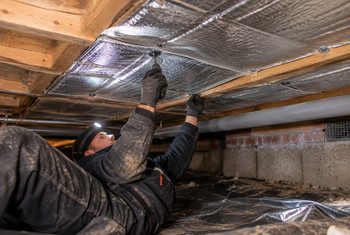 Monteur bevestigt reflecterende thermokussens tegen de onderzijde van een houten vloer in een kruipruimte